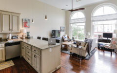 Modern open-concept living room and kitchen with arched windows, light cabinets, granite counters, hardwood floors, TV, and sofa.