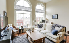 Bright living room with arched windows, white sofa, wooden table, TV, wicker chair, decor items; city buildings visible outside.