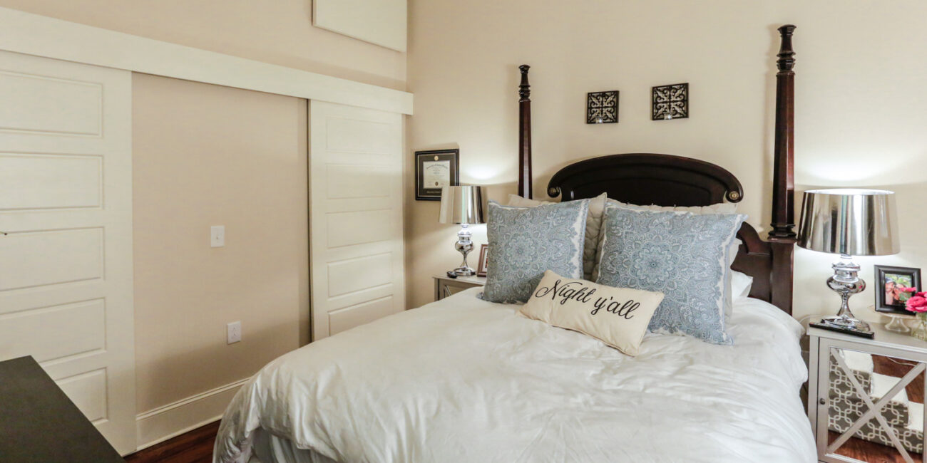 A cozy bedroom with a four-poster bed, white bedding, blue patterned pillows, and a Night y'all decorative pillow. Mirrored nightstands with lamps and a sliding closet door are in the background.