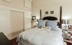 A cozy bedroom with a four-poster bed, white bedding, blue patterned pillows, and a Night y'all decorative pillow. Mirrored nightstands with lamps and a sliding closet door are in the background.