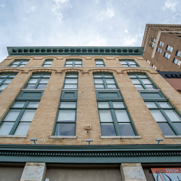 Low-angle view of a historic yellow brick building with green windows, decorative trim and partly cloudy sky; taller buildings flank both sides.
