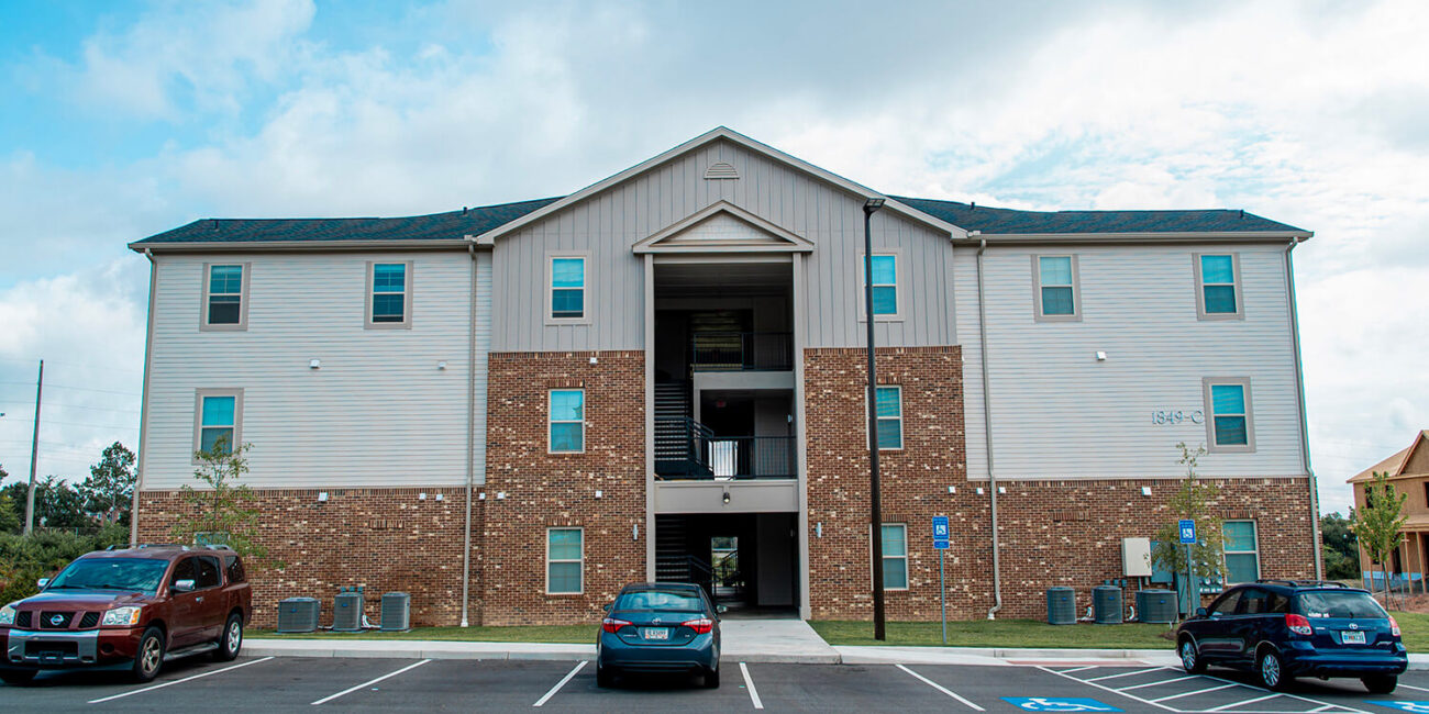 Three-story brick and light-sided apartment building, four cars out front, central entrance, mostly empty lot, partly cloudy sky.