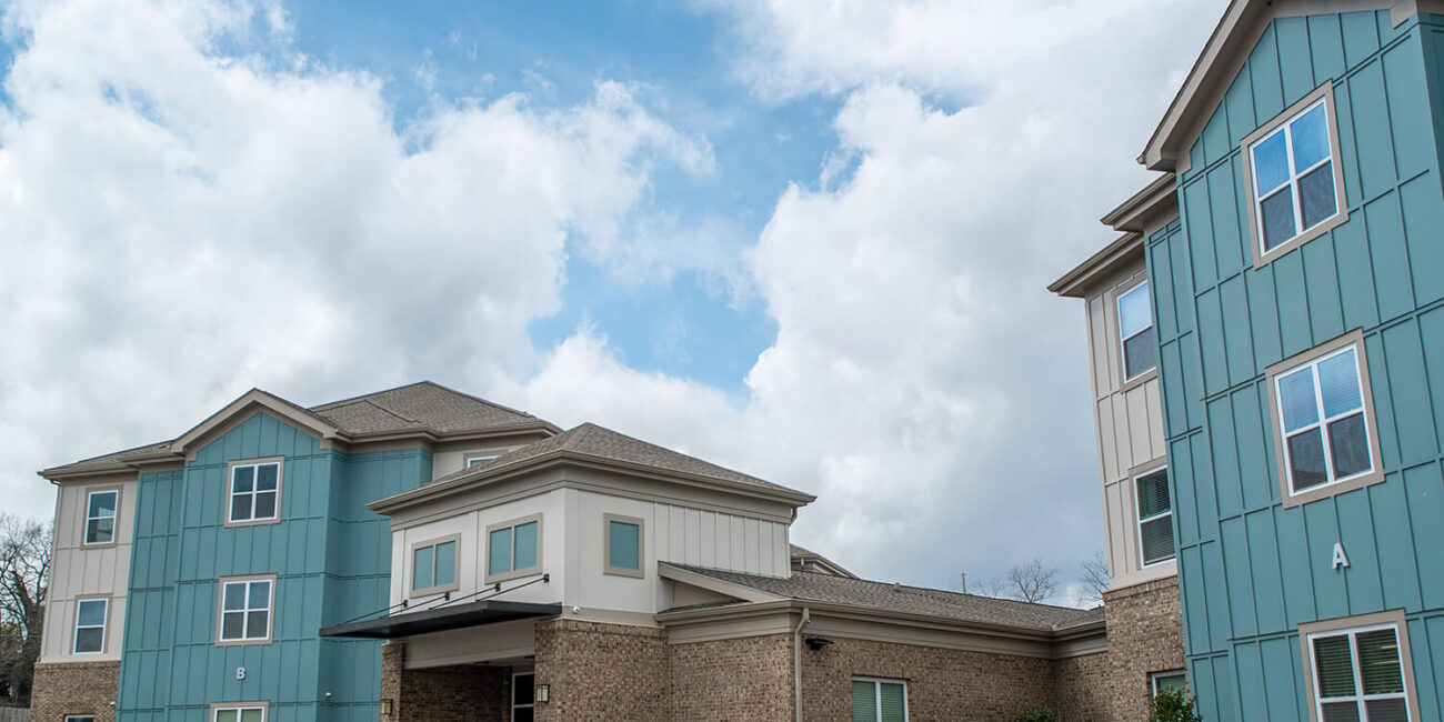 A modern apartment building with blue and beige siding, brick accents, and front landscaping, under a partly cloudy sky.
