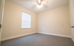 An empty room with beige walls, closed-blind window, gray carpet, white baseboard, outlets, and a ceiling fan with lights.