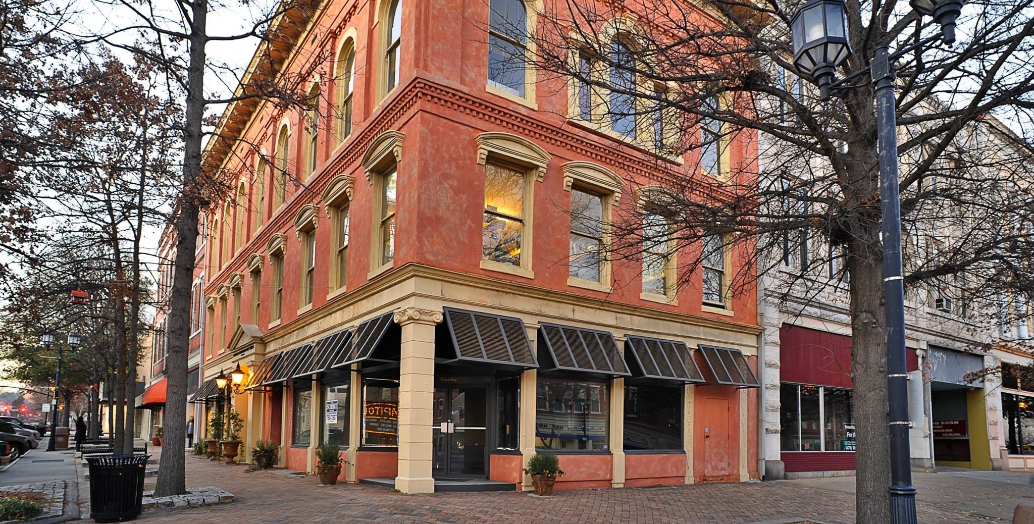 Three-story orange and tan building with large windows and black striped awnings on a tree-lined downtown street corner with brick sidewalks.