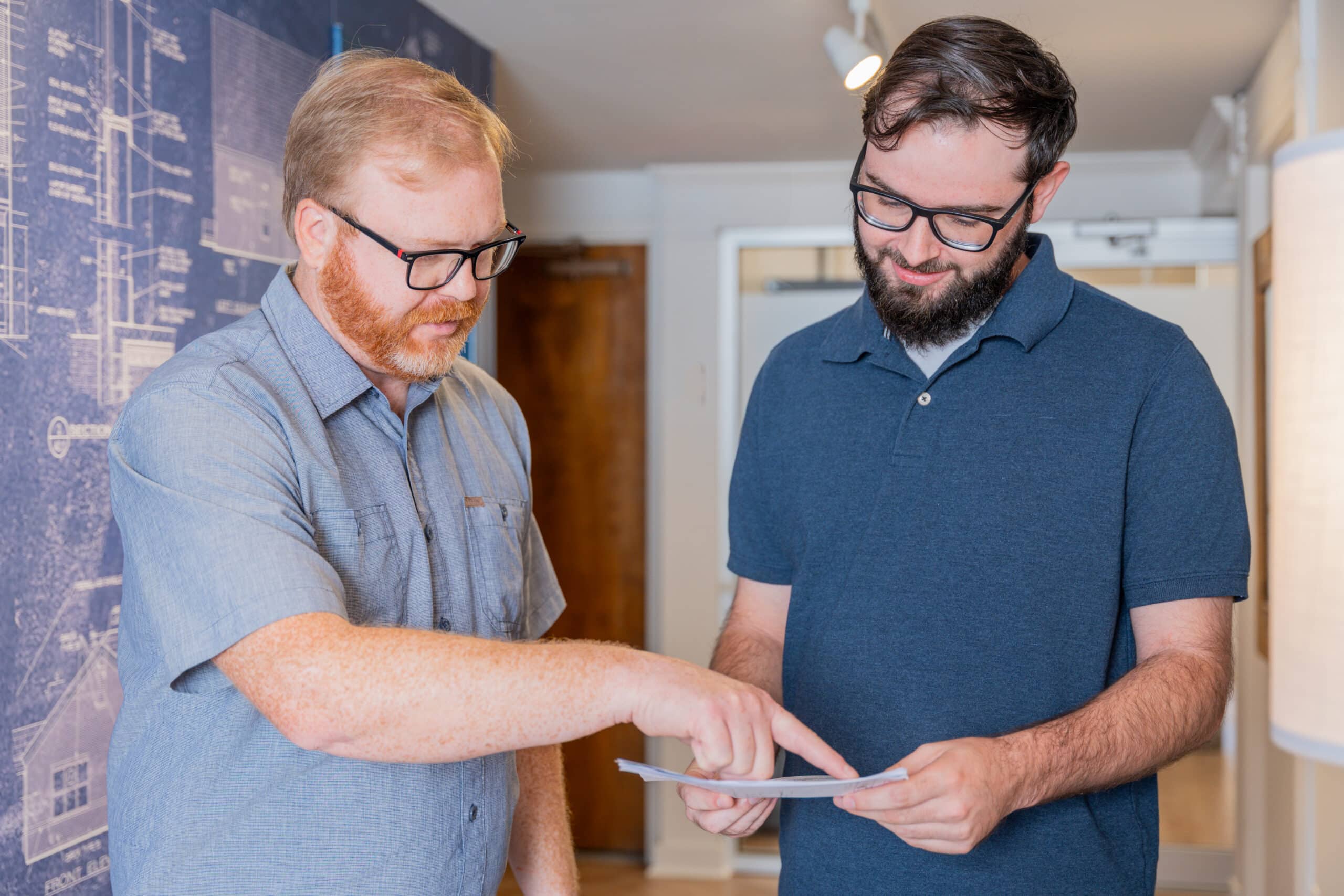 Two bearded men with glasses in short-sleeved shirts stand indoors, discussing a document—one points as the other holds it.
