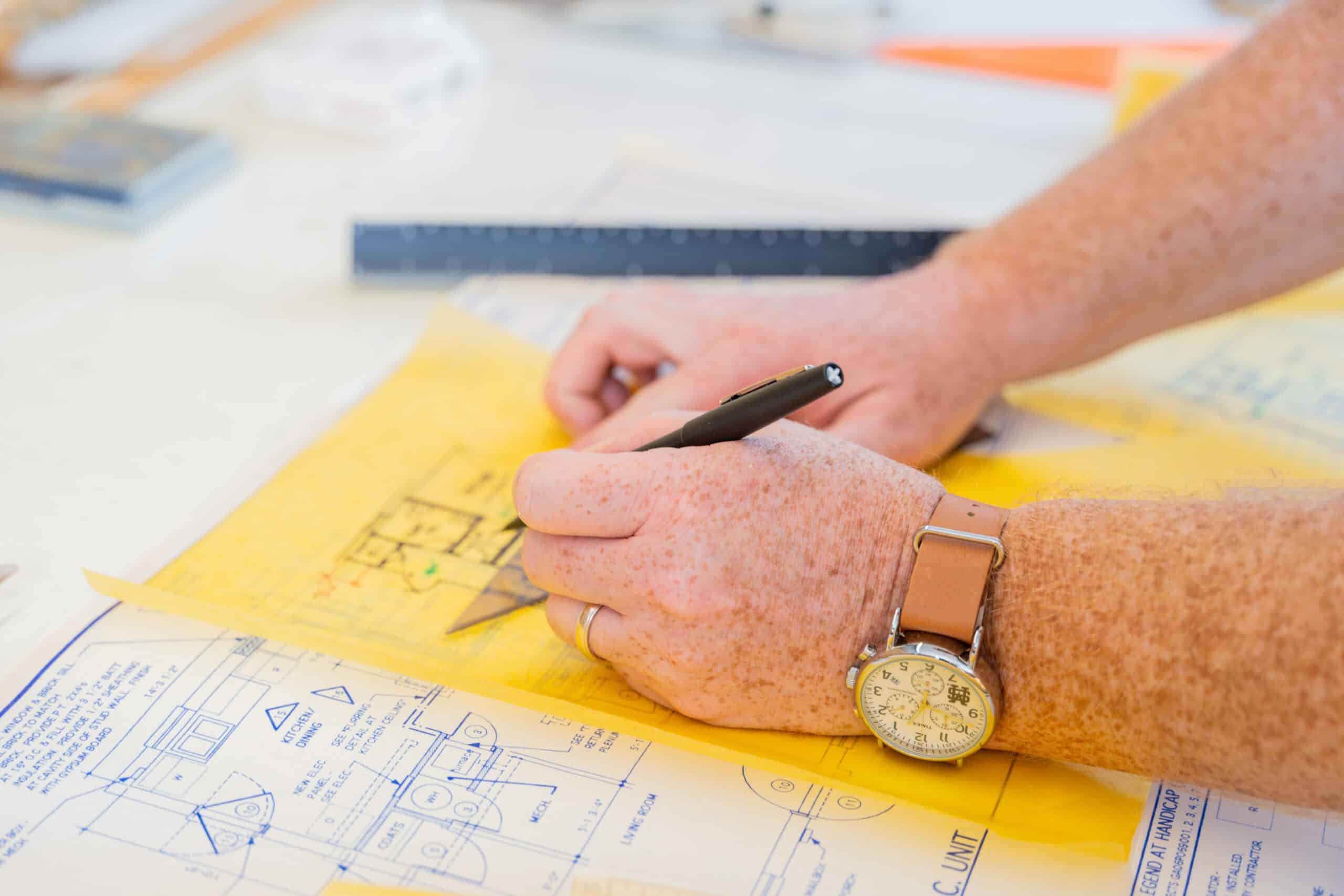 A person with a watch sketches on yellow graph paper over technical blueprints, using a black pen and ruler, drawings on the desk.