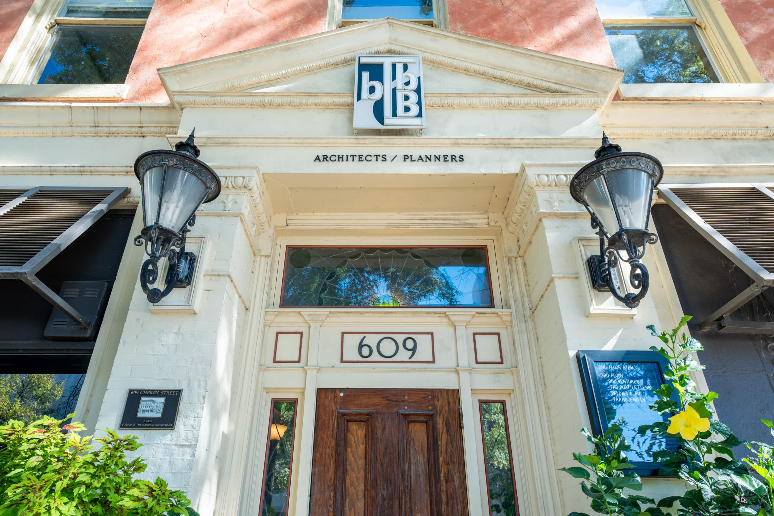 Entrance to a building at 609 with large glass lanterns, an Architects / Planners sign under a logo, and greenery right of the door.