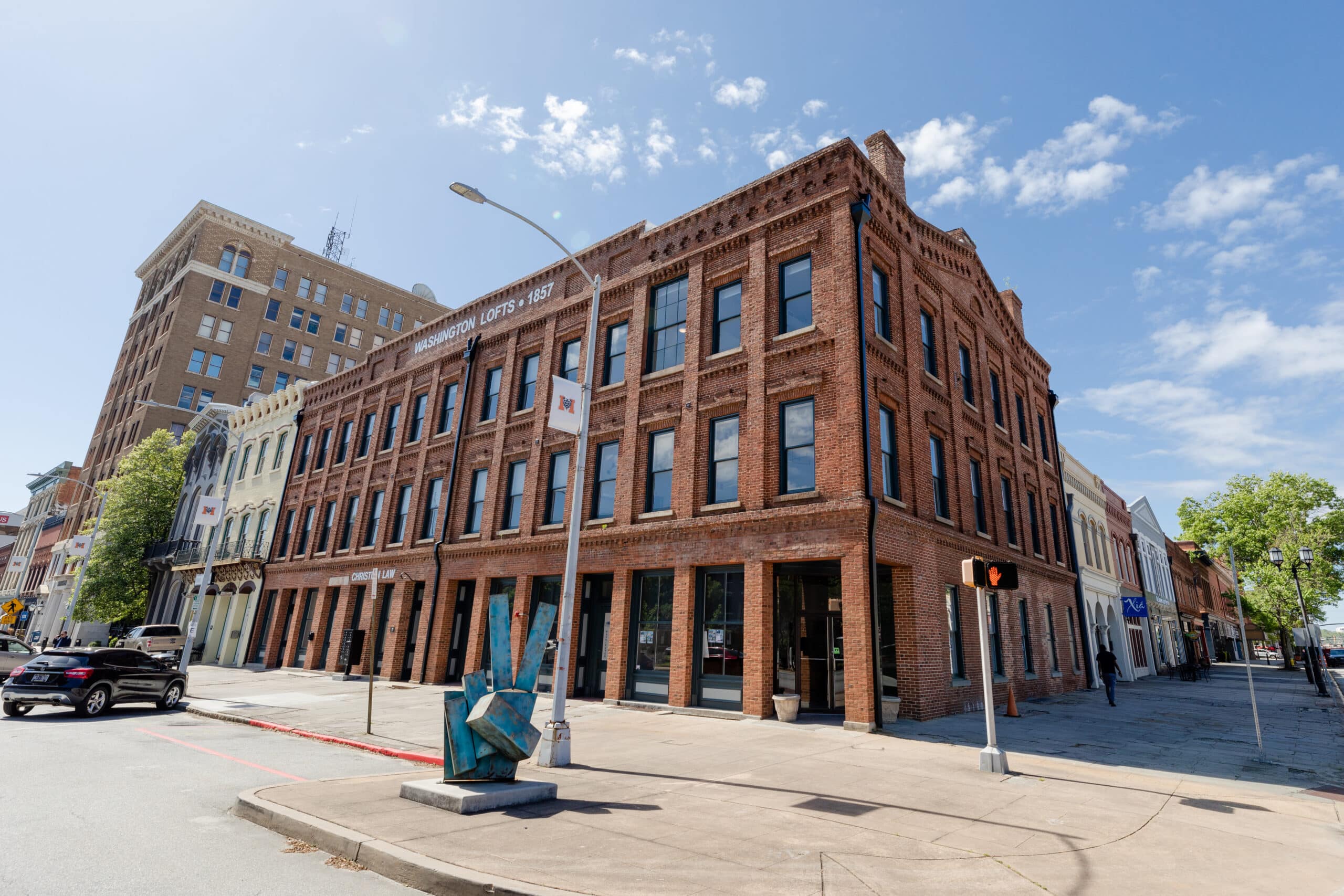 Historic three-story red brick building on a street corner under blue sky, a few parked cars, and a modern sculpture out front.