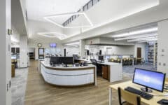 Modern library interior with computers on desks, curved help desk, bookshelves, seating areas, wood and carpet floors, large windows.