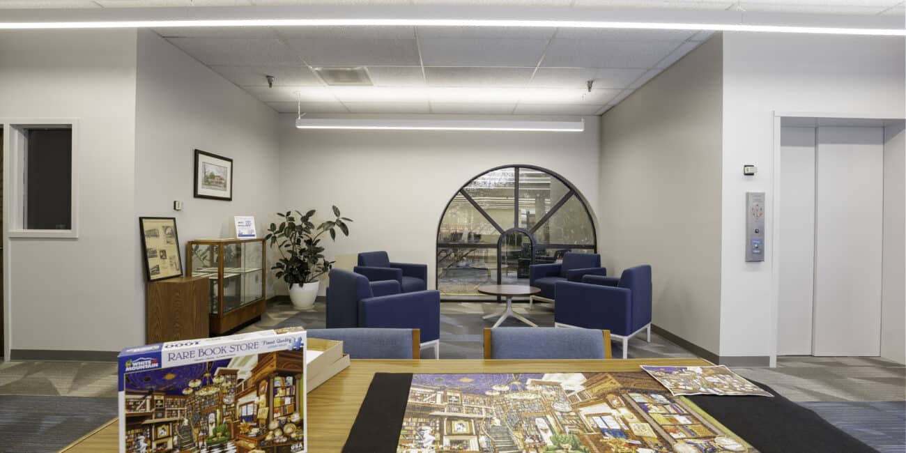 A bright, modern room with blue armchairs, a round window, glass display case, puzzle on a table, and elevator to the right.