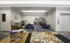A bright, modern room with blue armchairs, a round window, glass display case, puzzle on a table, and elevator to the right.