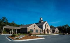 Modern church with stone and beige siding, arched entrance, steeple and cross, by a small lot under blue sky with shrubs and trees.