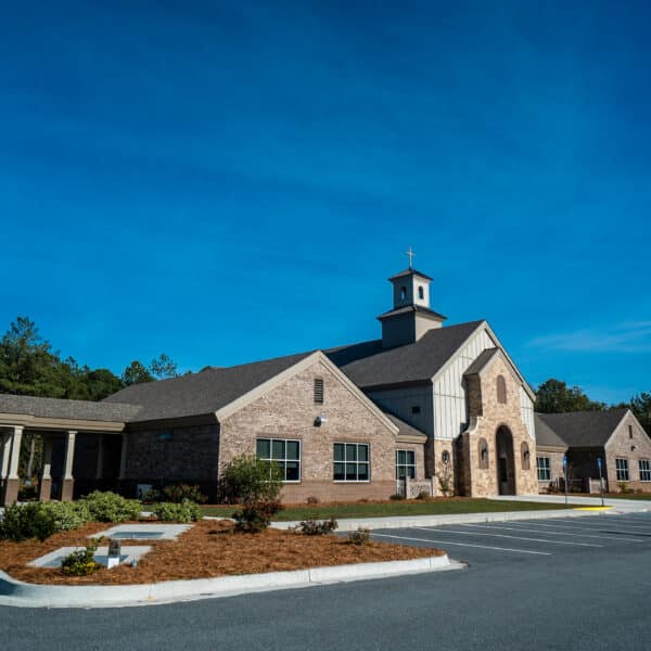 Modern church with stone and beige siding, arched entrance, steeple and cross, by a small lot under blue sky with shrubs and trees.