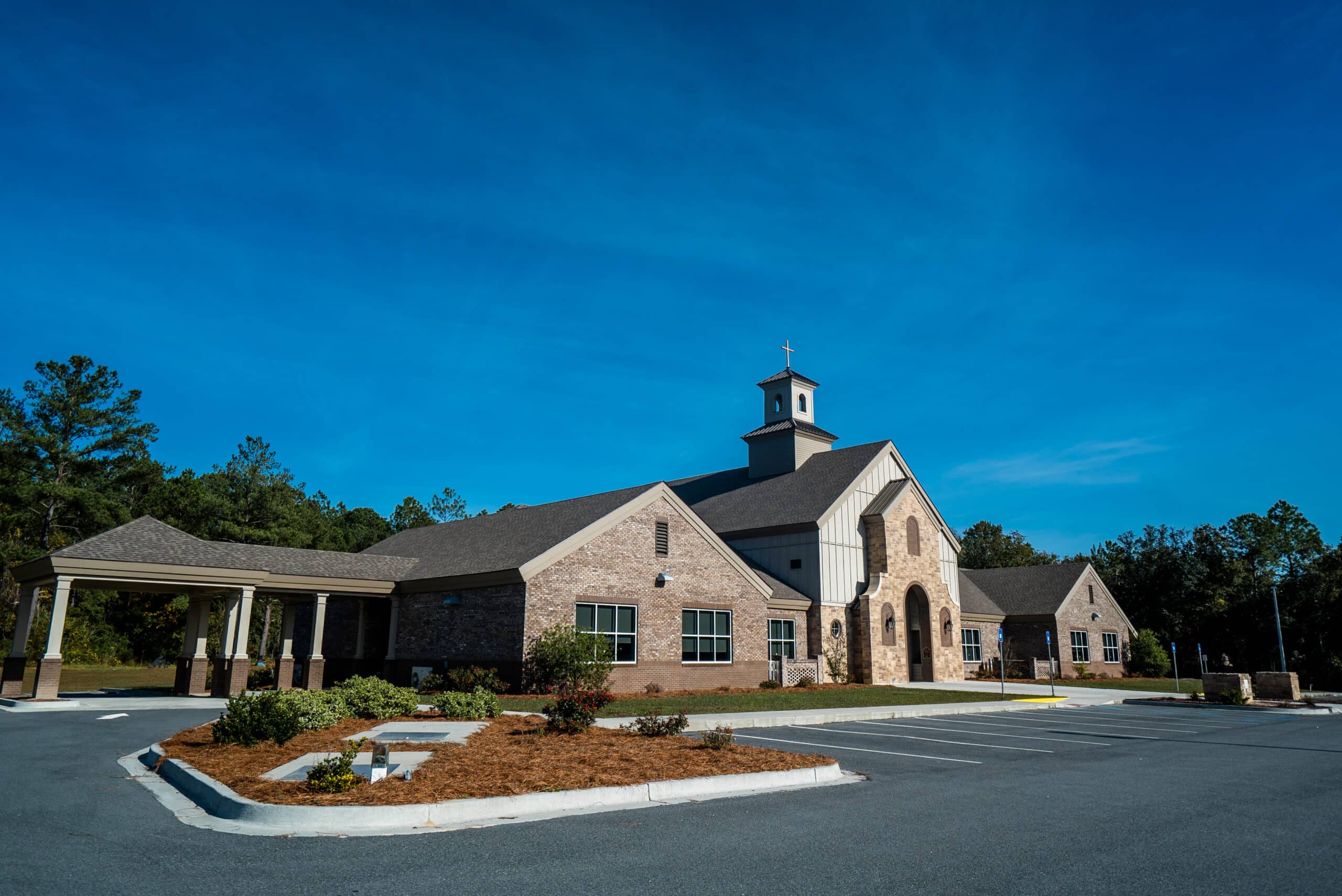 Modern church with stone and beige siding, arched entrance, steeple and cross, by a small lot under blue sky with shrubs and trees.
