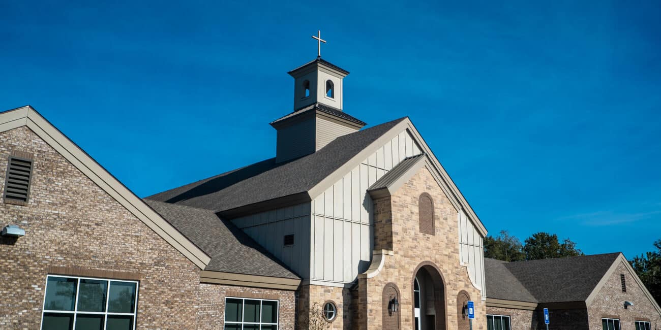 Modern brick church with large windows and a cross-topped steeple under a blue sky; accessible parking signs by the entrance.