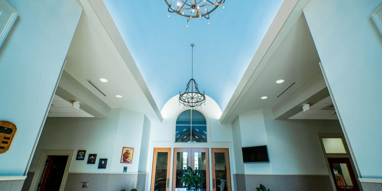 Modern lobby with vaulted ceilings, chandeliers, big windows, double wood doors, potted plants, wall art, and central table.