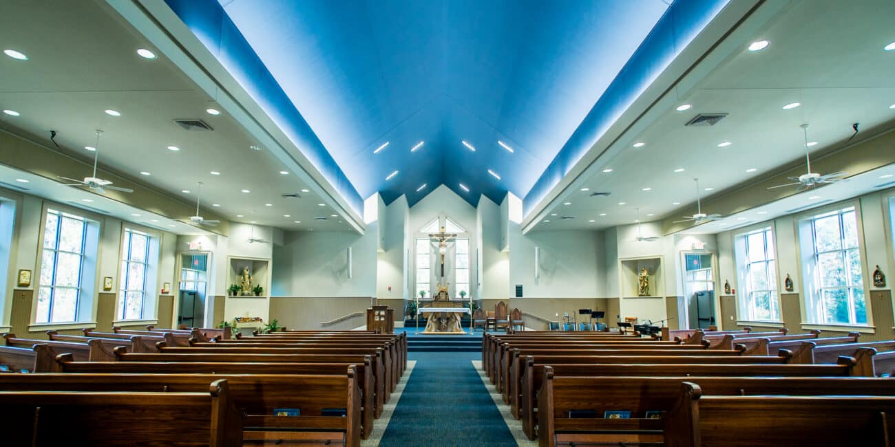 Bright modern church interior with wooden pews, blue ceiling, and large windows. Aisle leads to altar and cross. Natural light fills space.