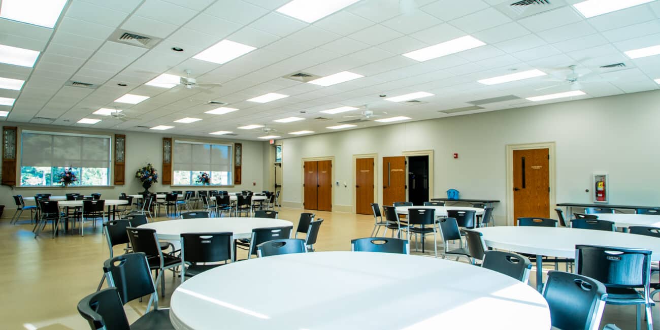 A bright, spacious room with round and rectangular tables, black chairs, large windows, light walls, and ceiling lights ready for an event.