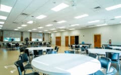 A bright, spacious room with round and rectangular tables, black chairs, large windows, light walls, and ceiling lights ready for an event.
