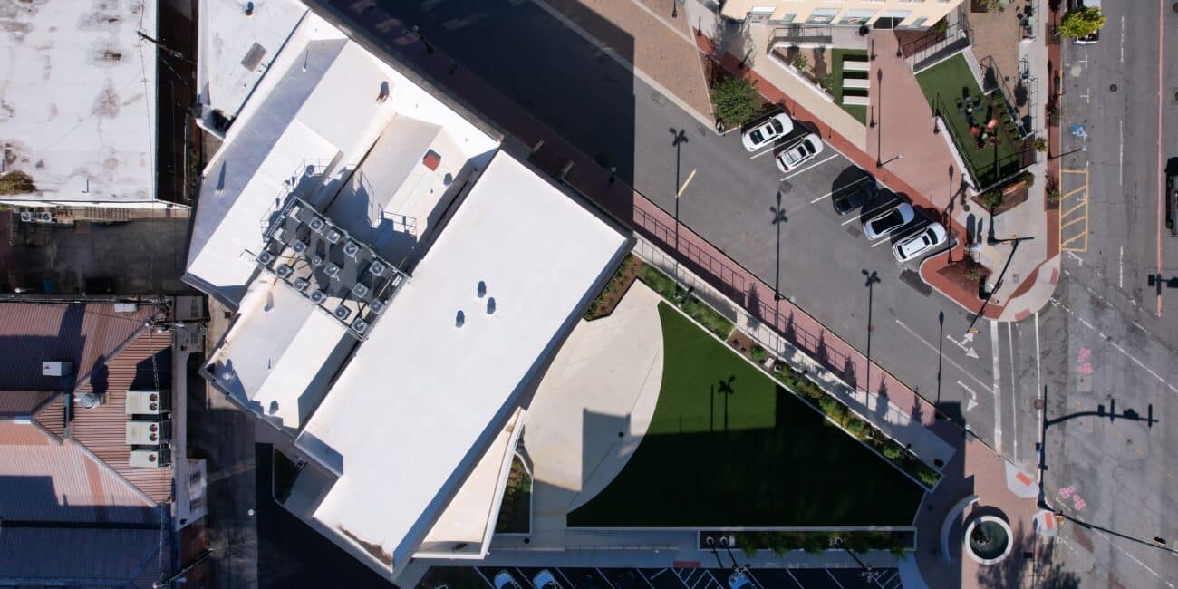 Aerial view of a modern building with white roof, green lawn, parking lots, and nearby city streets with cars and sidewalks.