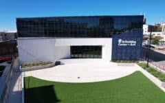 A modern building with large glass windows and a sign reading Otis Redding Center for the Arts stands behind a courtyard with green artificial turf and concrete paths under a clear blue sky.