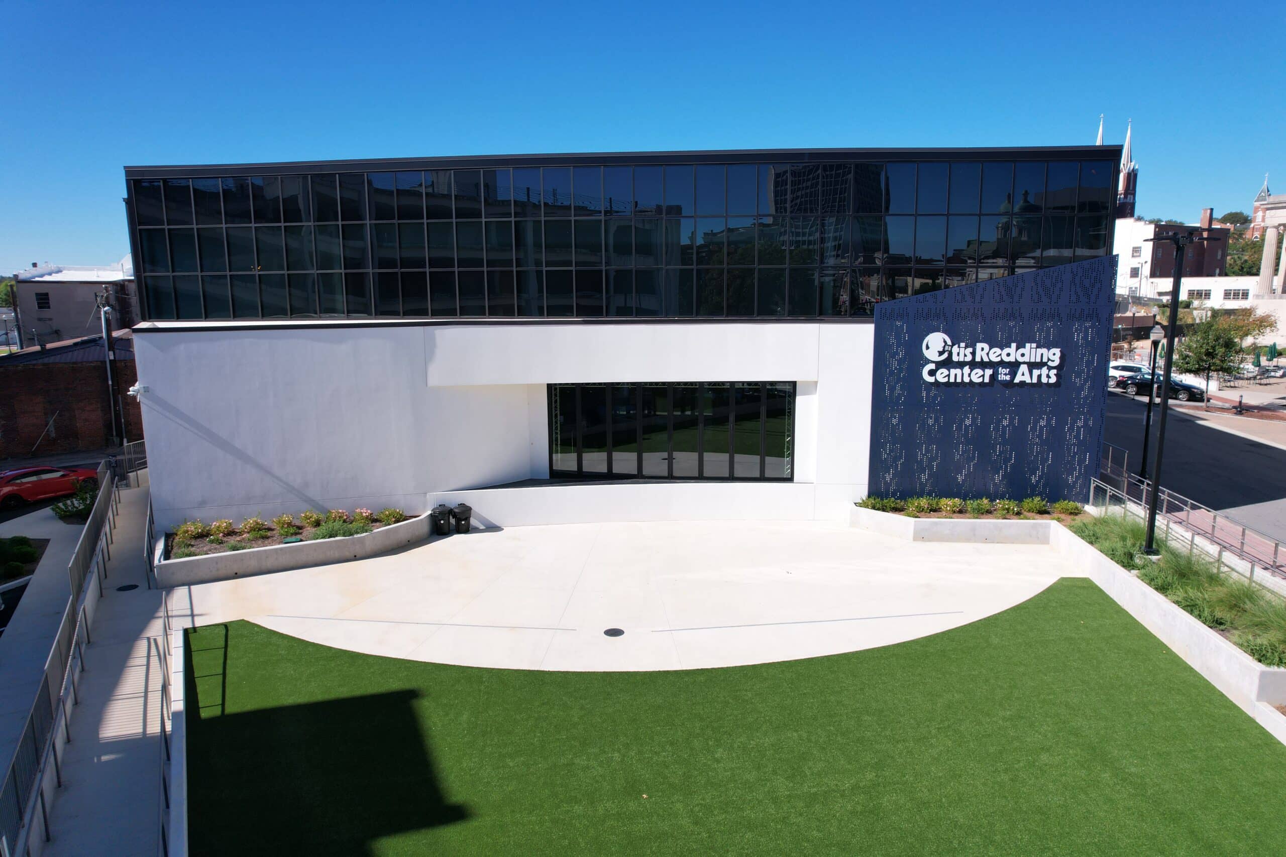 A modern building with large glass windows and a sign reading Otis Redding Center for the Arts stands behind a courtyard with green artificial turf and concrete paths under a clear blue sky.