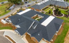 Aerial view of a modern brick building complex with dark gray roofs, green lawns, pathways, parking lots, and surrounding roads.