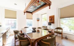 Bright meeting room with a glass-top table, eight green chairs, stained glass ceiling panel, large windows, and sunlight streaming in.