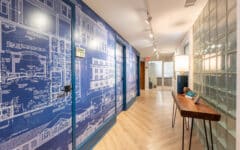 Modern hallway with light wood floors, a wall featuring big blue architectural blueprints, glass block windows, and track lighting.
