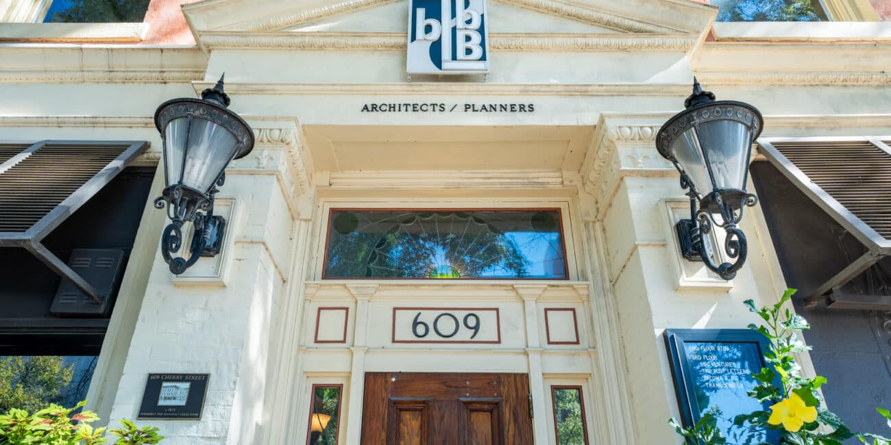 Entrance to a building with the number 609 above a wooden door, large decorative lamps on both sides, a sign reading Architects / Planners, and green foliage in the foreground.