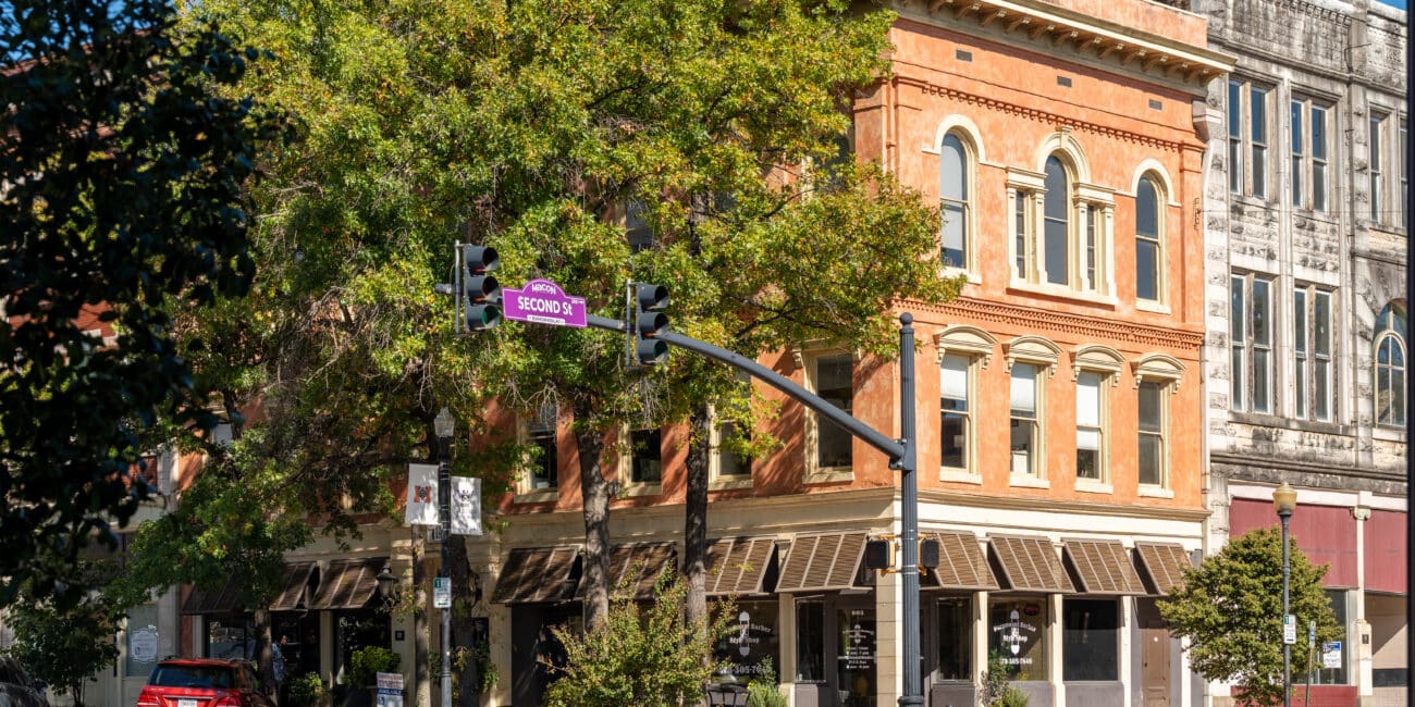A street corner with historic brick buildings, large windows, awnings, a red car by the curb, tree shade, and a purple Second St sign.