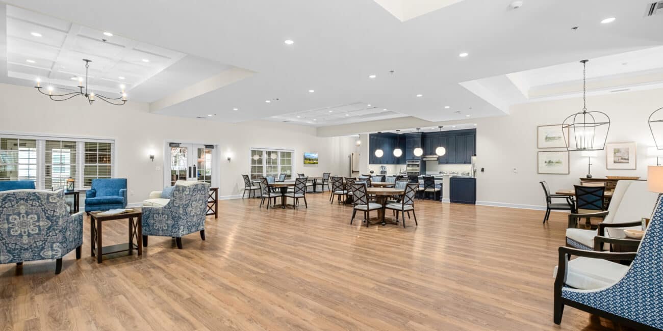 Spacious, well-lit dining and lounge area with wood floors, blue and beige armchairs, round tables, windows, and modern lighting.
