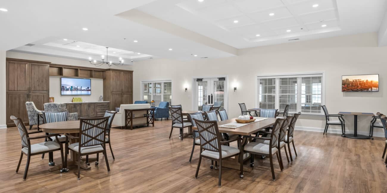 Spacious modern communal room with wood floors, tables, chairs, large TV, comfy seating. Well-lit with ceiling lights; welcoming vibe.