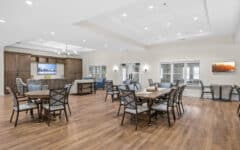 Spacious modern communal room with wood floors, tables, chairs, large TV, comfy seating. Well-lit with ceiling lights; welcoming vibe.