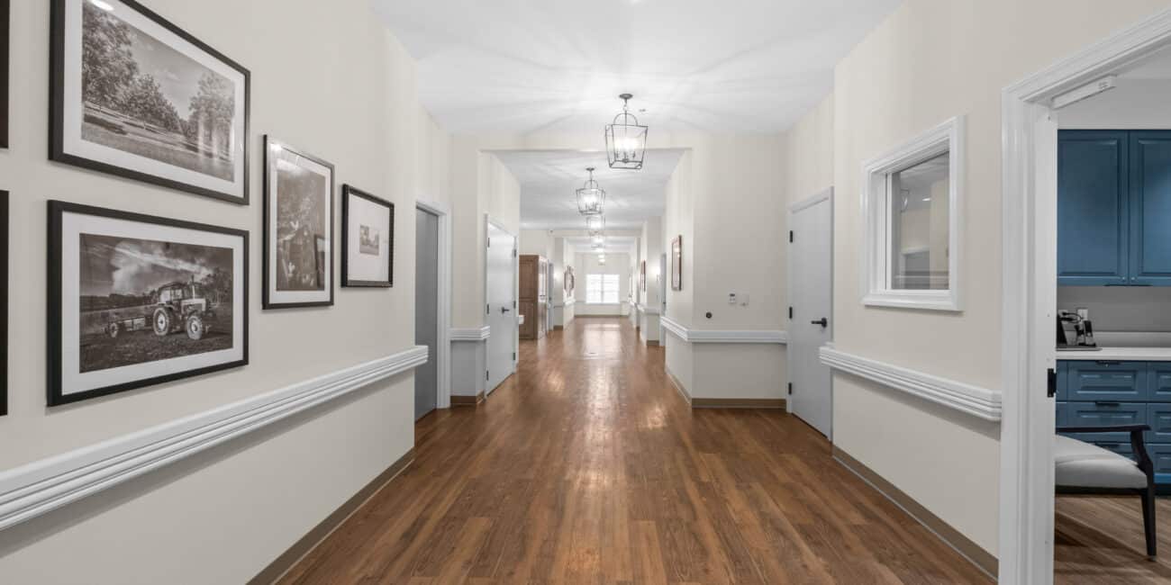 Well-lit hallway with wooden floors, cream walls, black-and-white photos on the left, and a blue office area seen through a window.