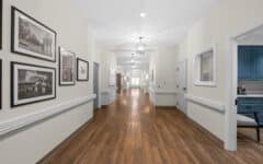 Well-lit hallway with wooden floors, cream walls, black-and-white photos on the left, and a blue office area seen through a window.