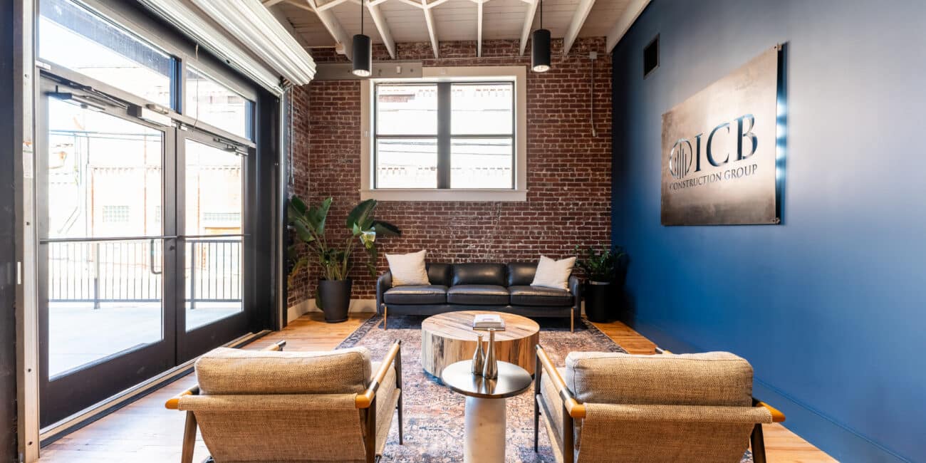 Modern office waiting area with two wooden chairs, a round coffee table, black sofa, plants, brick walls, big windows, JCB sign.