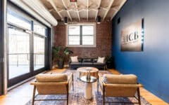 Modern office waiting area with two wooden chairs, a round coffee table, black sofa, plants, brick walls, big windows, JCB sign.