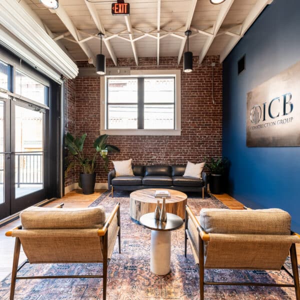 Modern office waiting area with two wooden chairs, a round coffee table, black sofa, plants, brick walls, big windows, JCB sign.