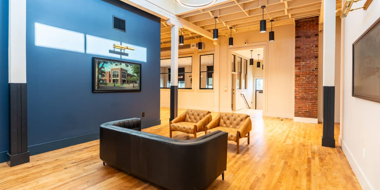 Modern, spacious room with wooden floors, blue accent wall, mounted TV, black curved sofa, tan armchairs, brick and large windows.