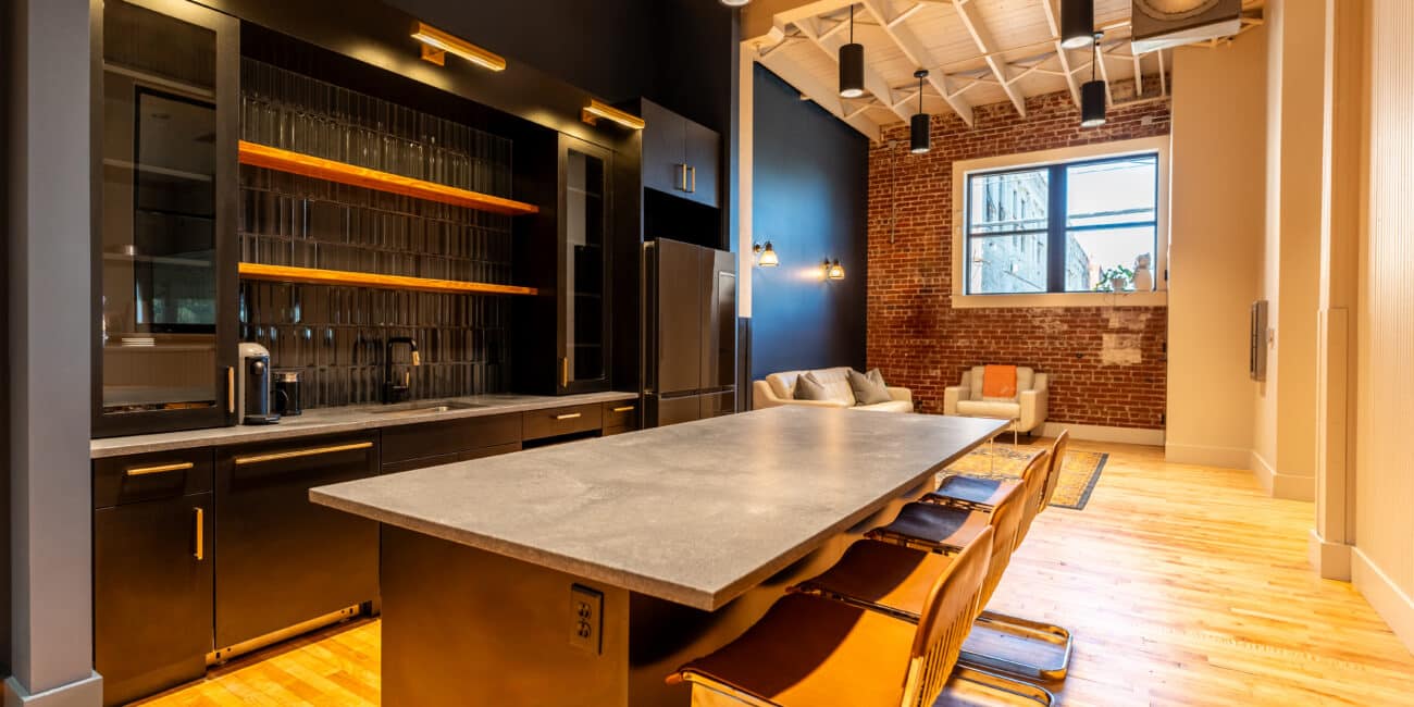 Modern kitchen featuring dark cabinets, open shelves, concrete island with four leather chairs, wood floors, and industrial accents.