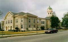 A beige two-story courthouse with a green-domed clock tower stands by a street where a maroon car is parked, surrounded by trees.