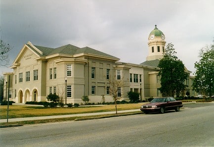 A beige two-story courthouse with a green-domed clock tower stands by a street where a maroon car is parked, surrounded by trees.
