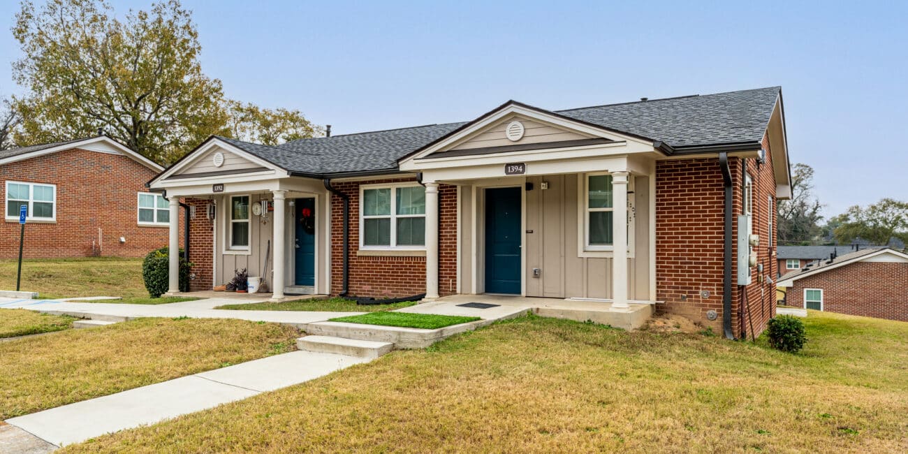 Single-story brick duplex with two blue doors, small porches, concrete walkways, grassy yard, and a few trees in the background.