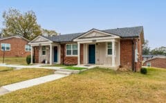 Single-story brick duplex with two blue doors, small porches, concrete walkways, grassy yard, and a few trees in the background.