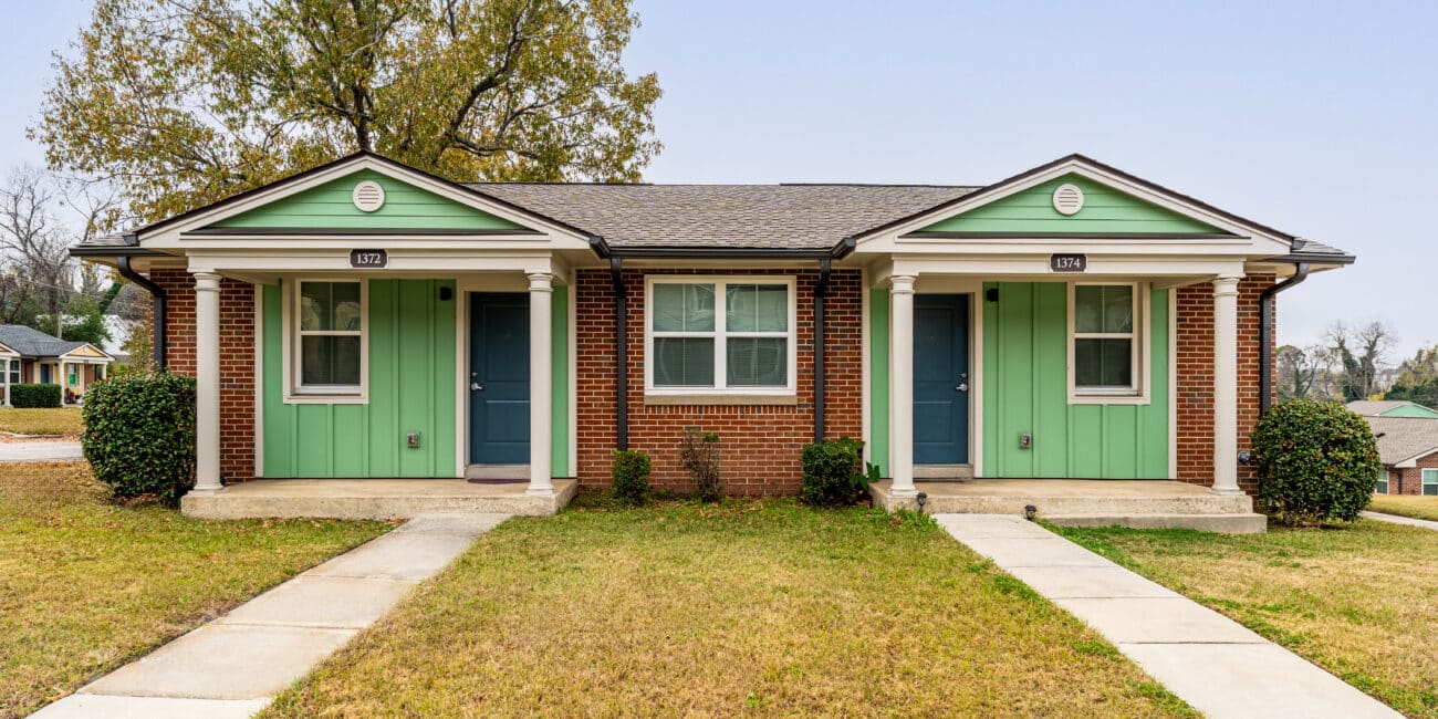 A duplex house with red brick walls, green and white siding, blue doors, a big tree behind, two walkways, grass, clear sky.