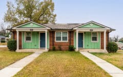 A duplex house with red brick walls, green and white siding, blue doors, a big tree behind, two walkways, grass, clear sky.