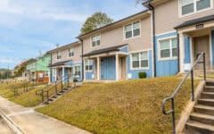 Modern two-story townhouses with colorful siding, front porches, and steps from a sloped yard beside a quiet sidewalk on a clear day.
