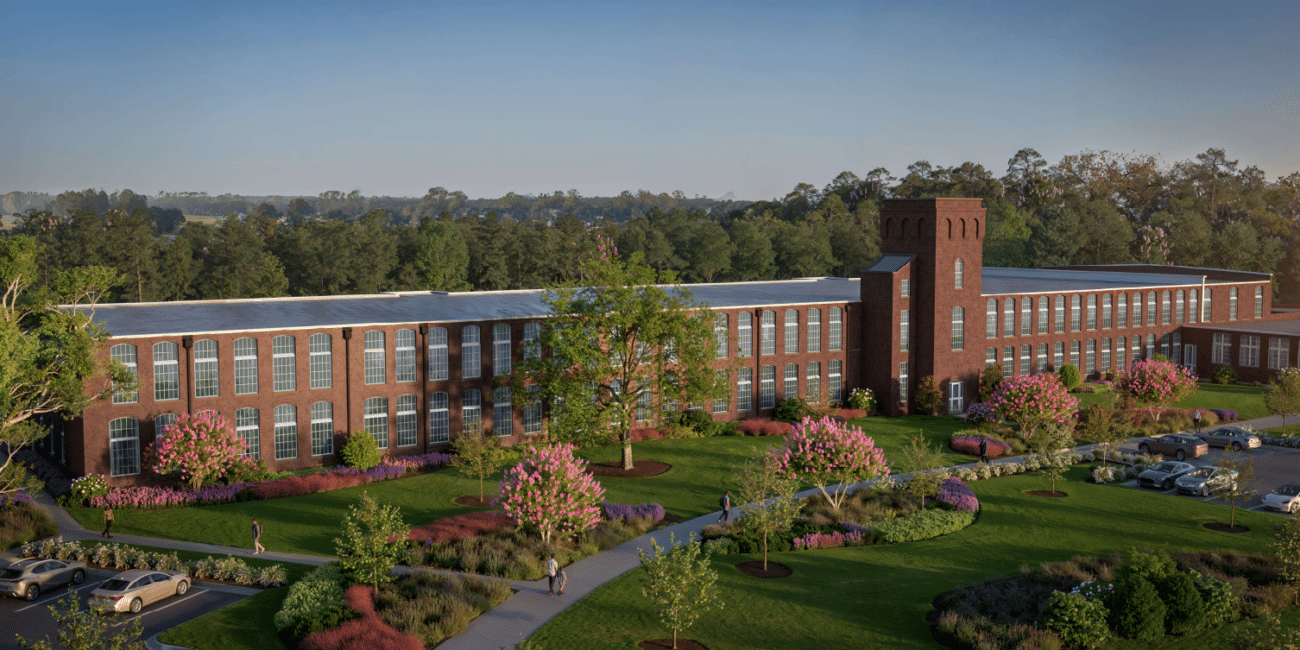 A red-brick building with tall windows and a central tower stands in lush gardens. Cars park nearby, trees and blue sky behind.